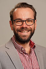 Smiling headshot of Mike Regilio wearing a grey suit jacket and red checkered collard shirt